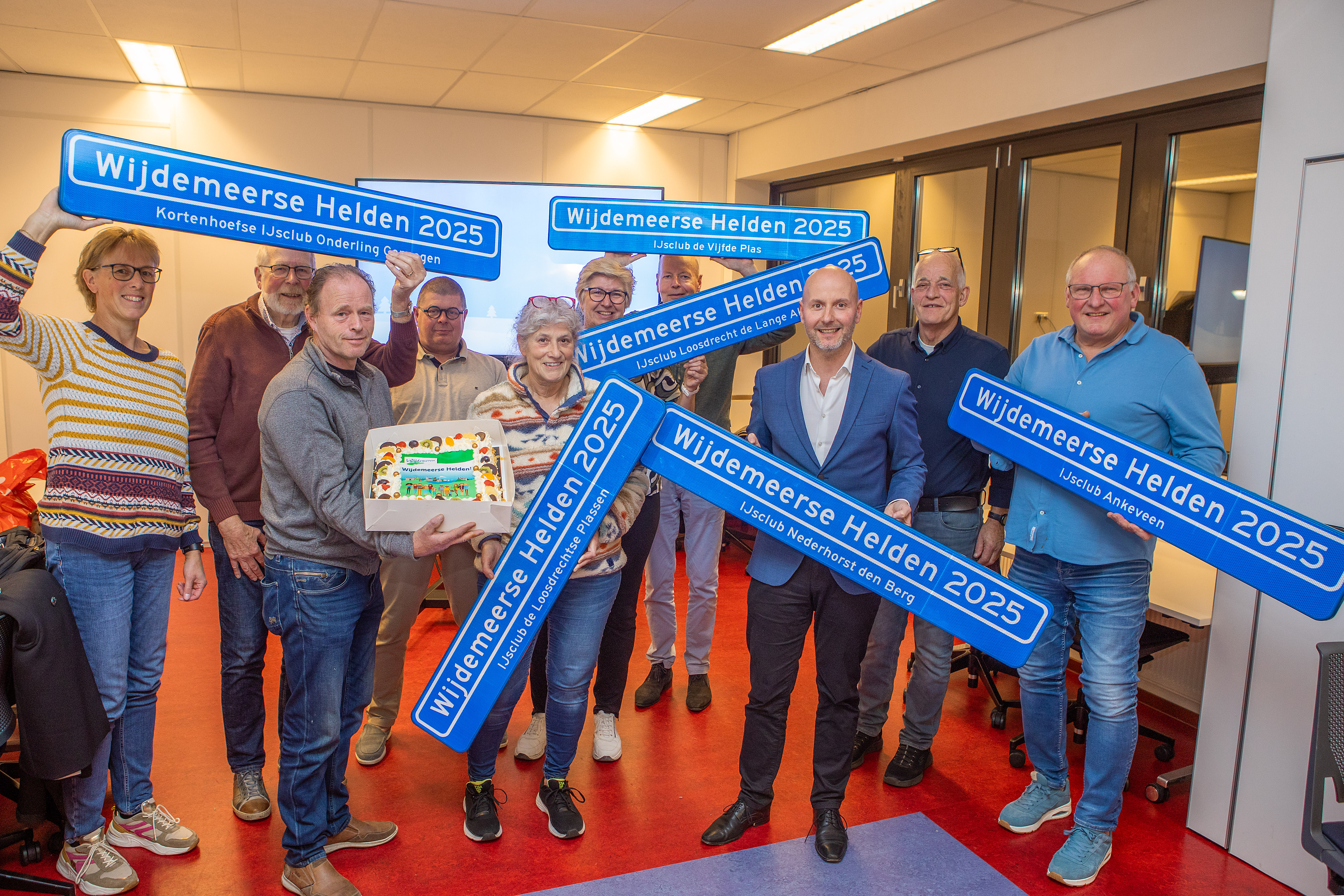 Op de foto zien we leden van de verschillende ijsclubs in Wijdemeren met burgemeester Mark Verheijen en taart. De vrijwilligers houden blauwe straatnaamborden vast met daarop de tekst 